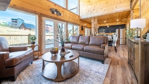 A spacious living room with a large window, a leather sofa, a coffee table, and a kitchen area with stainless steel appliances. - Take It Easy (Sevierville)