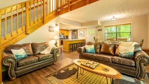 A living room with two leather sofas, a wooden staircase, a kitchen in the background, and a large window. - Bear Run Getaway (Gatlinburg)