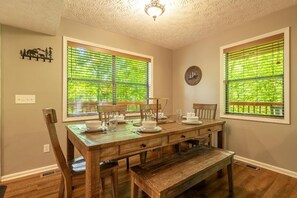 Ein Esszimmer mit Holztisch, Stühlen und einer Bank. Zwei Fenster mit Jalousien, eine Deckenleuchte und Wanddekorationen sind vorhanden.