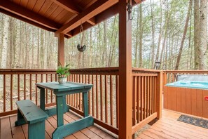 A wooden deck with a table and bench, a hot tub, and a view of a forest. - Starwood (Sevierville)