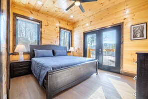 A wooden cabin bedroom with a bed, two bedside tables, a ceiling fan, and a large window. - Trinity (Sevierville)