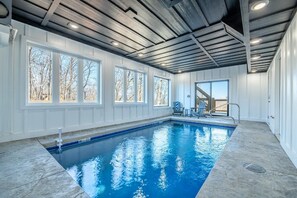 An indoor swimming pool with a view of trees through large windows, a ceiling with exposed beams, and a poolside area with chairs. - Trinity (Sevierville)