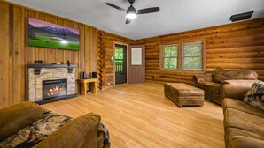 Ein Blockhaus-Interieur mit Kamin, Ledersofas, Deckenventilator und einem großen Fernseher an der Wand.