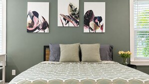 A bedroom with a bed, two pillows, a nightstand, a wall-mounted painting, and a window with blinds.