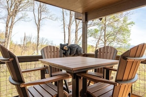 Eine Holzterrasse mit Tisch und Stühlen, einer Bärenskulptur und Blick auf Bäume sowie ein Gebäude im Hintergrund.