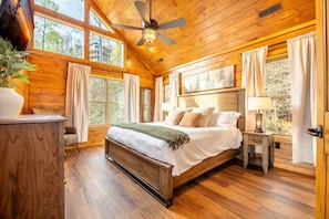 Ein Schlafzimmer einer Holzhütte mit einem Bett, Vorhängen, einem Deckenventilator und einem Fenster.