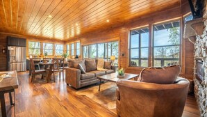 A spacious living area with wooden floors, a stone fireplace, and large windows offering a view of the outdoors. - Sunset Above the Smokies (Gatlinburg)
