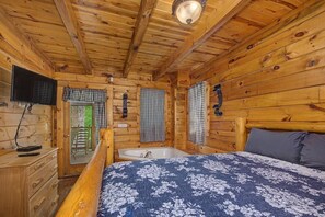 A wooden cabin room with a bed, a dresser, a television, and a window with a checkered curtain. - Sweet Fantasy (Sevierville)