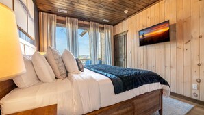 Un dormitorio con paneles de madera, una cama, cortinas, un televisor montado y una ventana.