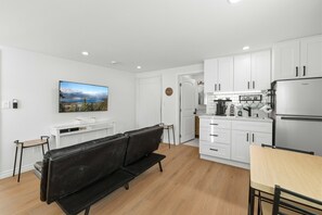 Eine moderne Wohnung mit Wohnbereich, ausgestattet mit einem schwarzen Ledersofa, einem Holzesstisch und einer Küche mit weißen Schränken sowie einem Edelstahlkühlschrank.