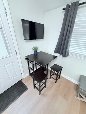 A small room with a dark wooden table, two matching chairs, a small potted plant, a mounted TV, and a window with grey curtains. - Luxury 3BR Retreat | Hot Tub, Fire Pit & Games (Simi Valley)