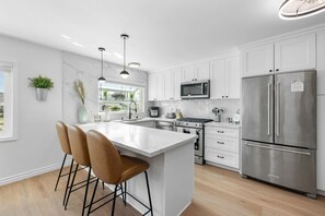 A modern kitchen with white cabinetry, a stainless steel refrigerator, a microwave, an oven, and a large island with bar stools. - Luxury 3BR Retreat | Hot Tub, Fire Pit & Games (Simi Valley)