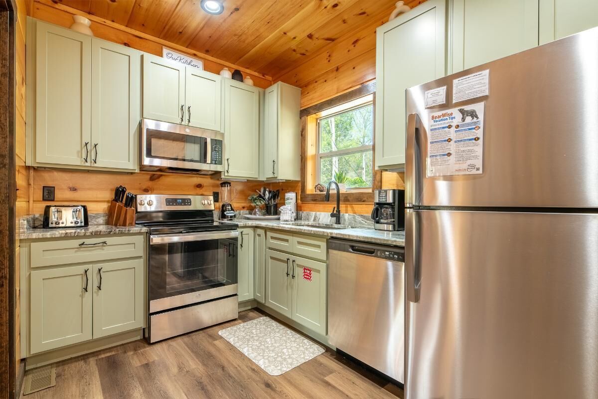 A kitchen with light green cabinets, stainless steel appliances, and a window with a view of trees.