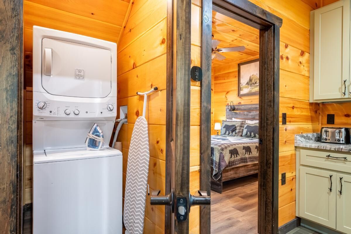 A compact room with a washing machine, a bed with a bear-themed bedspread, a small kitchenette, and wooden walls.