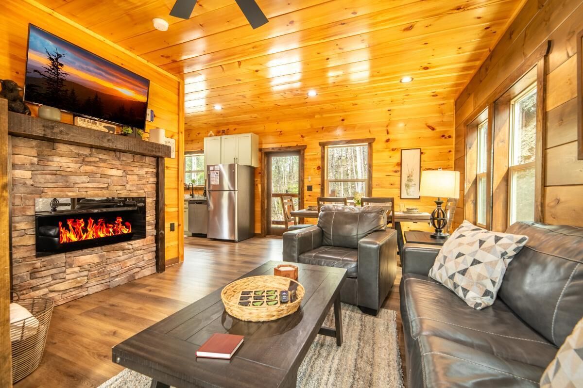 A log cabin interior with a stone fireplace, leather seating, a dining area, and a kitchen.