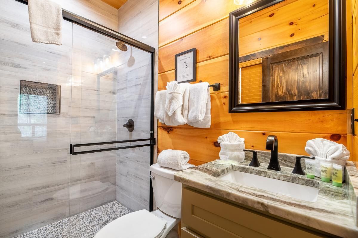 A bathroom with a glass shower, a toilet, a sink with a marble countertop, and wooden walls.