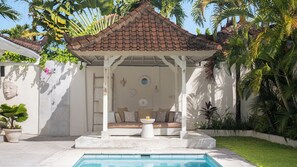 A covered outdoor seating area with a pool, surrounded by lush greenery and a decorative wall. - Villa Mas Estate (Kuta)