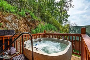 A hot tub on a wooden deck with a view of a rocky hillside and trees. - Z - Smoky Serenity Retreat (Sevierville)