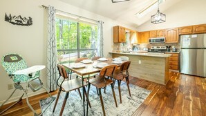 Uma sala de jantar com uma mesa posta para quatro pessoas, uma cadeirinha de alimentação, armários de madeira, uma geladeira de aço inoxidável e uma janela com cortinas.