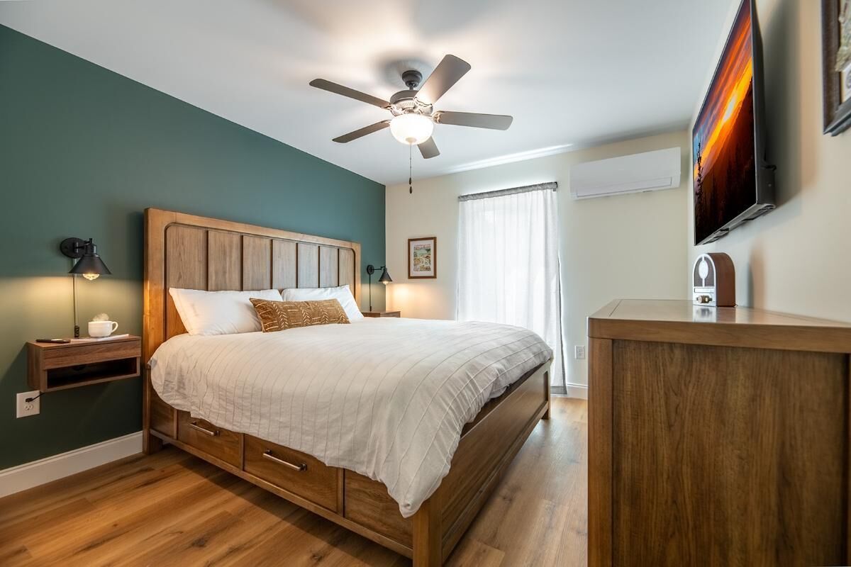 A bedroom with a wooden bed, a ceiling fan, a bedside table with a lamp, a TV mounted on the wall, and a window with curtains.