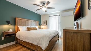 A bedroom with a wooden bed, a ceiling fan, a bedside table with a lamp, a TV mounted on the wall, and a window with curtains.