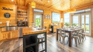 Interior de una cabaña de madera con cocina, comedor y amplias ventanas.