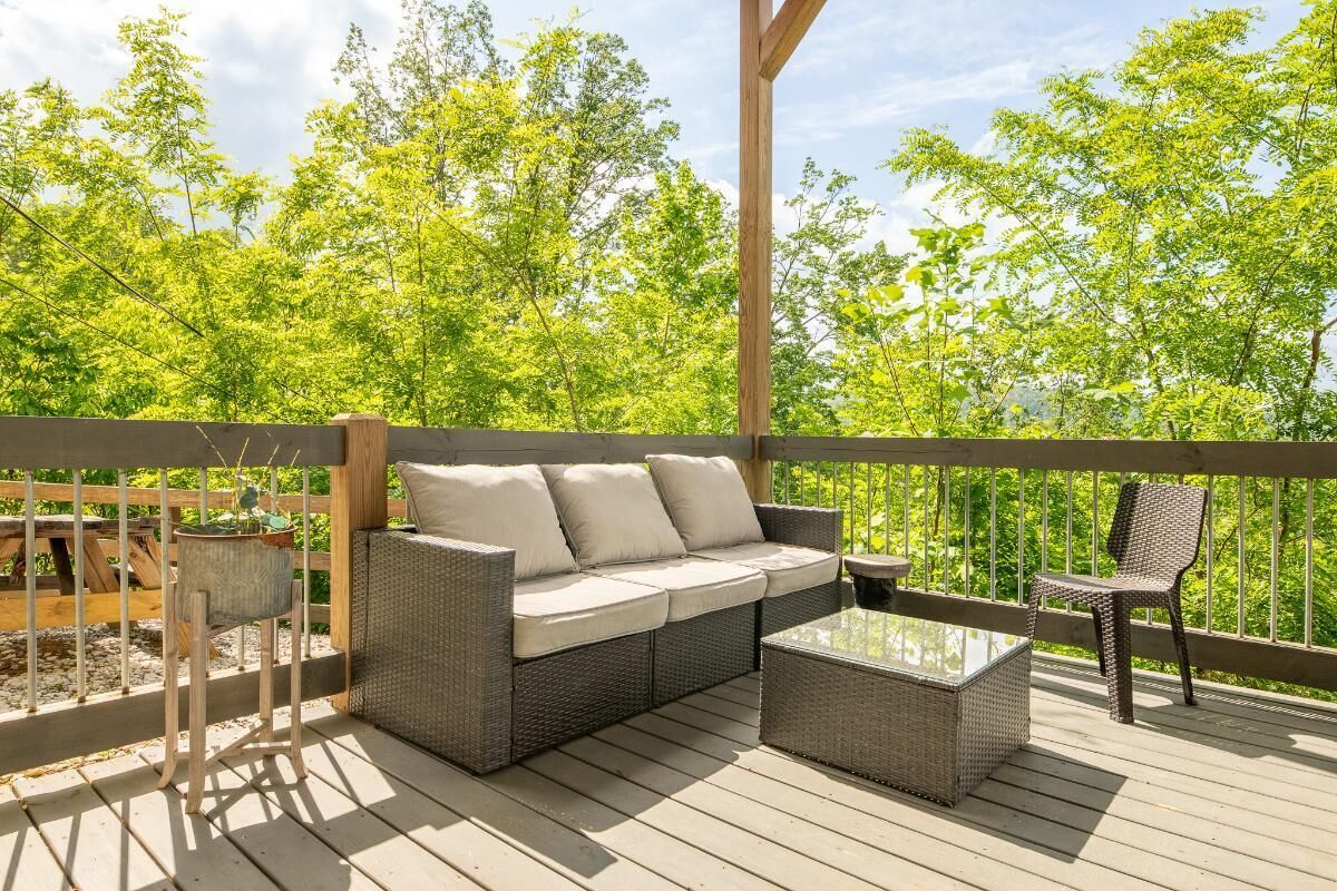 A covered outdoor seating area with a wicker sofa, a small table, and a chair, surrounded by a wooden deck and a metal railing.