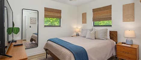 Une chambre à coucher comprenant un lit, des tables de chevet, un ventilateur de plafond, un miroir, une fenêtre avec des stores et une vue sur la verdure à l’extérieur.