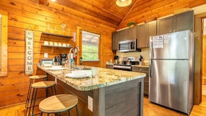 A wooden cabin interior with a kitchen featuring a central island, bar stools, a stainless steel refrigerator, and a microwave. - Lofty Pines (Sevierville)
