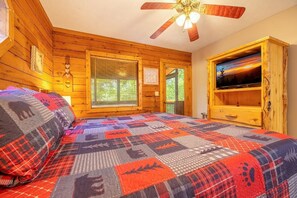 A wooden cabin bedroom with a bed, a ceiling fan, a TV on a wooden stand, and a window with blinds. - Lofty Pines (Sevierville)