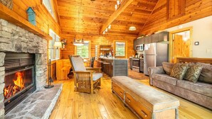 A log cabin interior with a stone fireplace, wooden furniture, a kitchen area, and a cozy seating arrangement. - Lofty Pines (Sevierville)