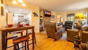 A living room with a fireplace, a dining area with a wooden table and chairs, and a kitchen in the background. - The Bears Den (Pigeon Forge)