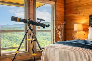 Ein Holzhüttenraum mit einem Teleskop auf einem Stativ, einem Bett mit blauer Decke und Blick auf einen Wald durch große Fenster.