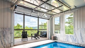En swimmingpool med ett glastak, utomhusmöbler och stora fönster som ger utsikt över omgivningen.