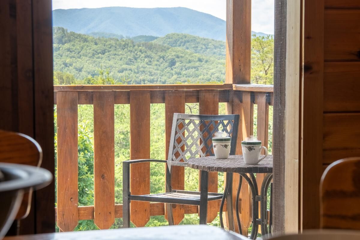 Terraza de madera con mesa y sillas, con vistas a una cadena montañosa boscosa.