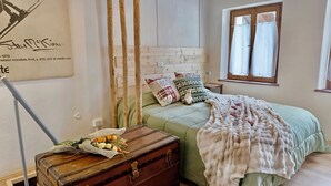 Ein Schlafzimmer mit einem Bett, einem Holzkopfende, einem Fenster mit Vorhängen, einem Nachttisch, einer Lampe und einem Koffer mit Blumen.