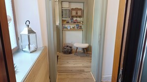 Ein schmaler Flur mit Holzboden, einem Korbwannenstuhl, einem kleinen Hocker und einem Regal mit Büchern und Dekorationsgegenständen.
