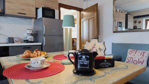 Eine Küche mit Mikrowelle, Kühlschrank und Kaffeemaschine. Ein Tisch mit Teekanne, Tassen und Gebäck ist zu sehen. Im Hintergrund befinden sich eine blaue Couch und ein dekoratives Kissen.
