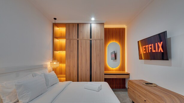 Una habitación de hotel moderna con una cama bien hecha, un armario de madera, una mesita de noche con una lámpara y un televisor de pared mostrando el logotipo de Netflix.