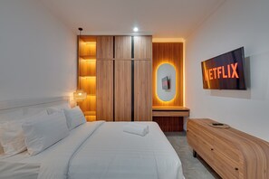 Una habitación de hotel moderna con una cama bien hecha, un armario de madera, una mesita de noche con una lámpara y un televisor de pared mostrando el logotipo de Netflix.