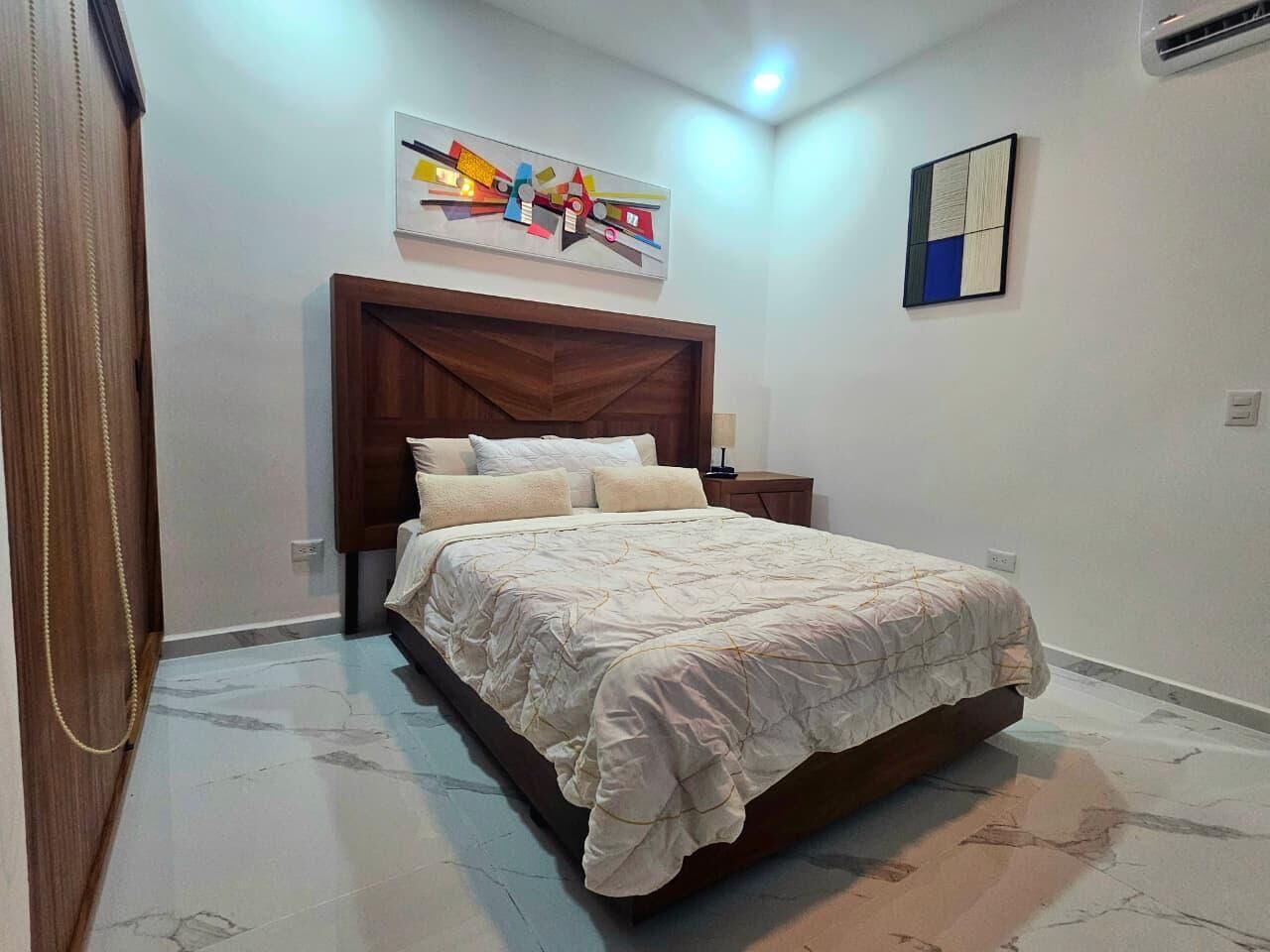A bedroom with a bed, a wooden headboard, a wall-mounted abstract art piece, a small bedside table, and a wall-mounted light fixture.