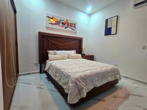 A bedroom with a bed, a wooden headboard, a wall-mounted abstract art piece, a small bedside table, and a wall-mounted light fixture. - Huayacan Cancún Apartment #2 (Cancún)