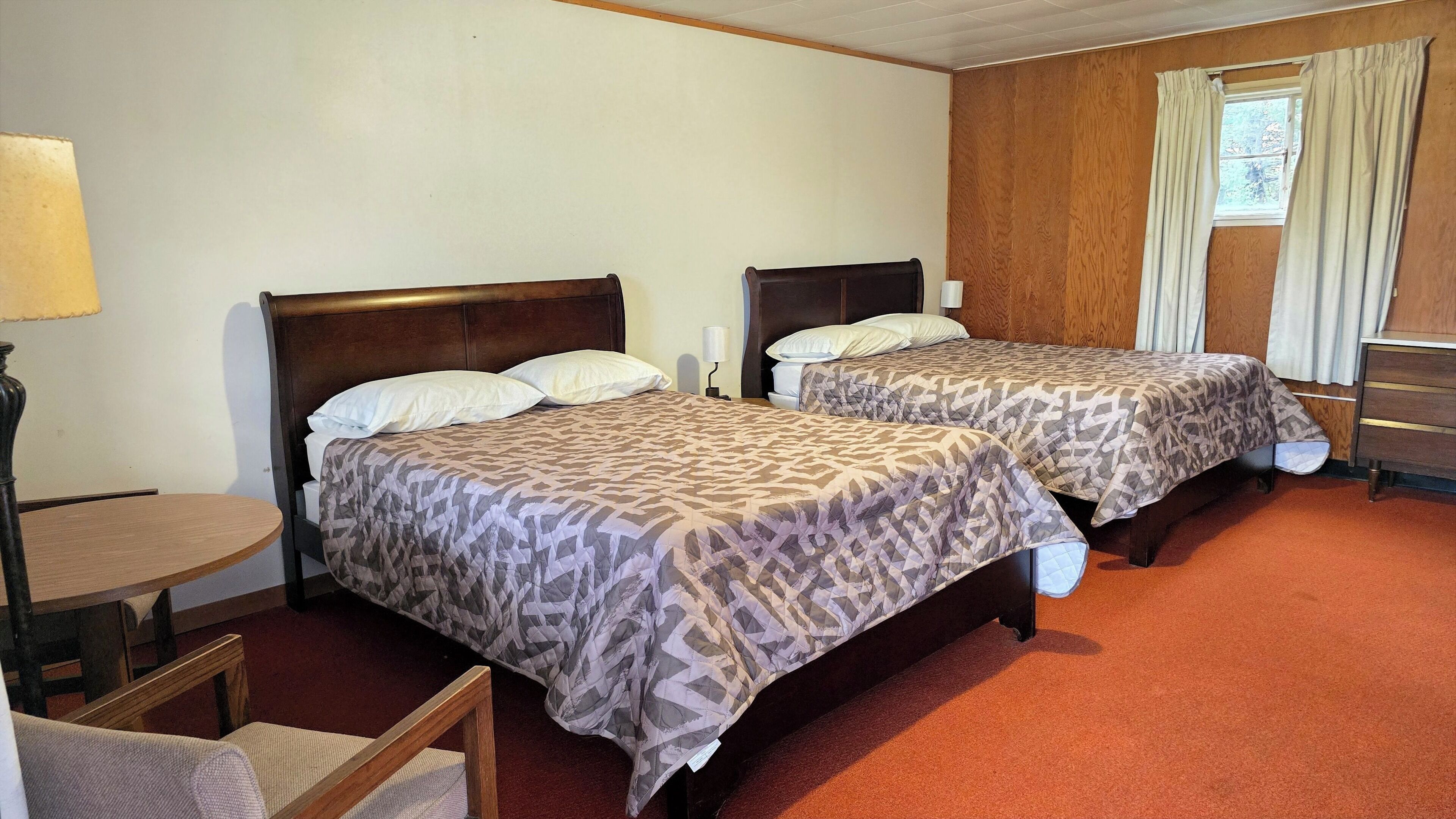 Ein Hotelzimmer mit zwei Betten, einem kleinen Tisch, einem Stuhl und einem Fenster mit Vorhängen.