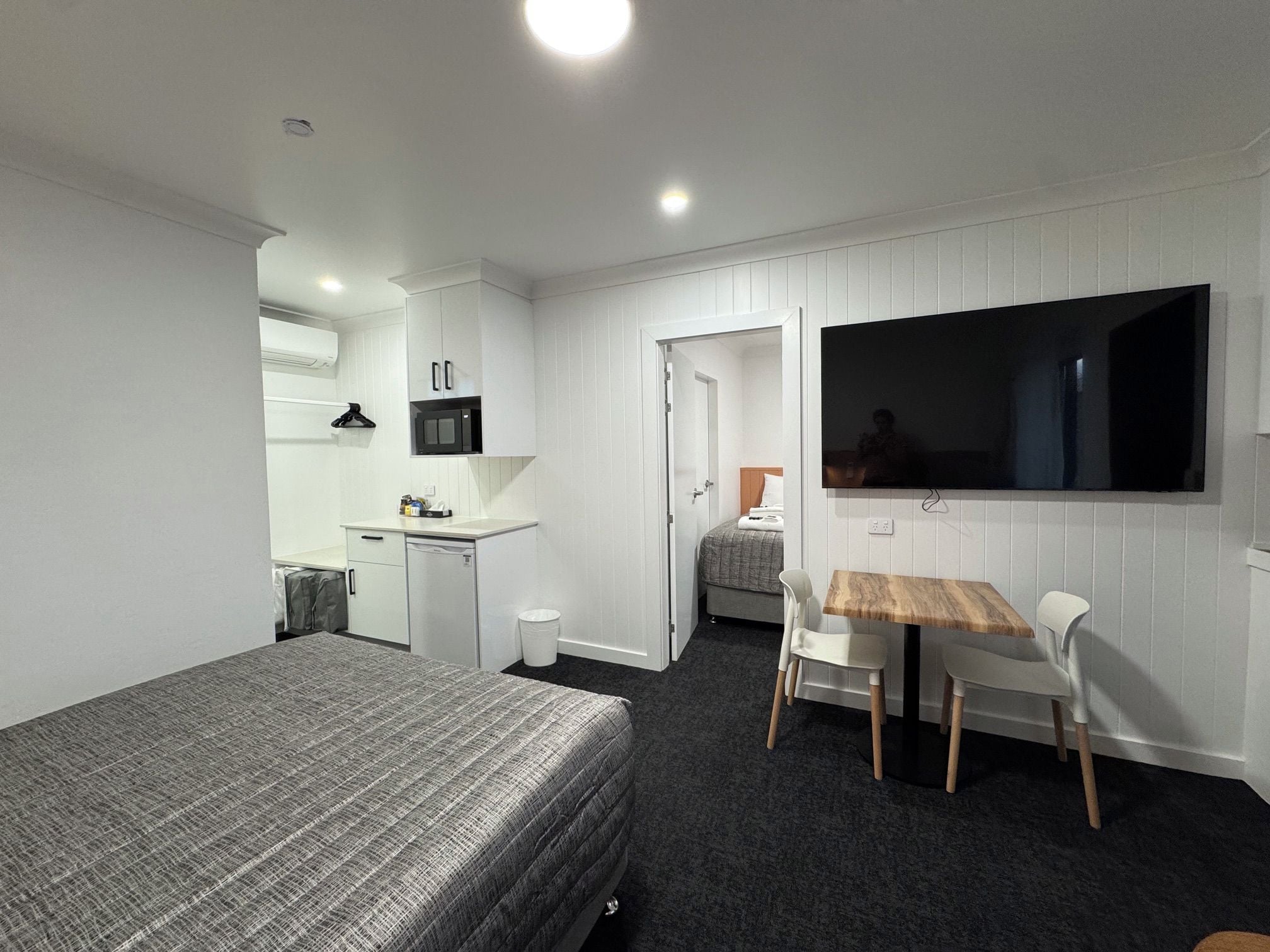 Una habitación funcional con cama, una pequeña kitchenette, un comedor con mesa y sillas, y un televisor fijado a la pared.