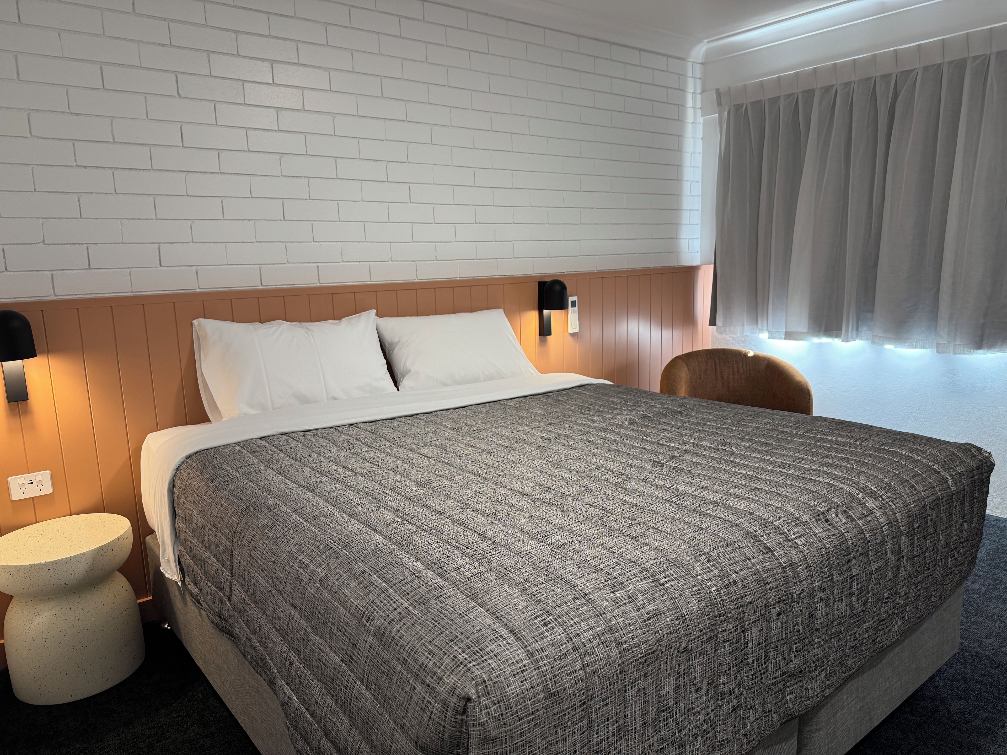 Habitación de hotel con una cama grande, una mesita redonda, un cabecero de madera y una ventana con cortinas.