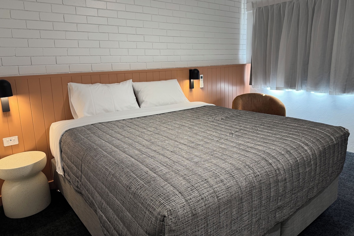 Habitación de hotel con una cama grande, una mesita redonda, un cabecero de madera y una ventana con cortinas.