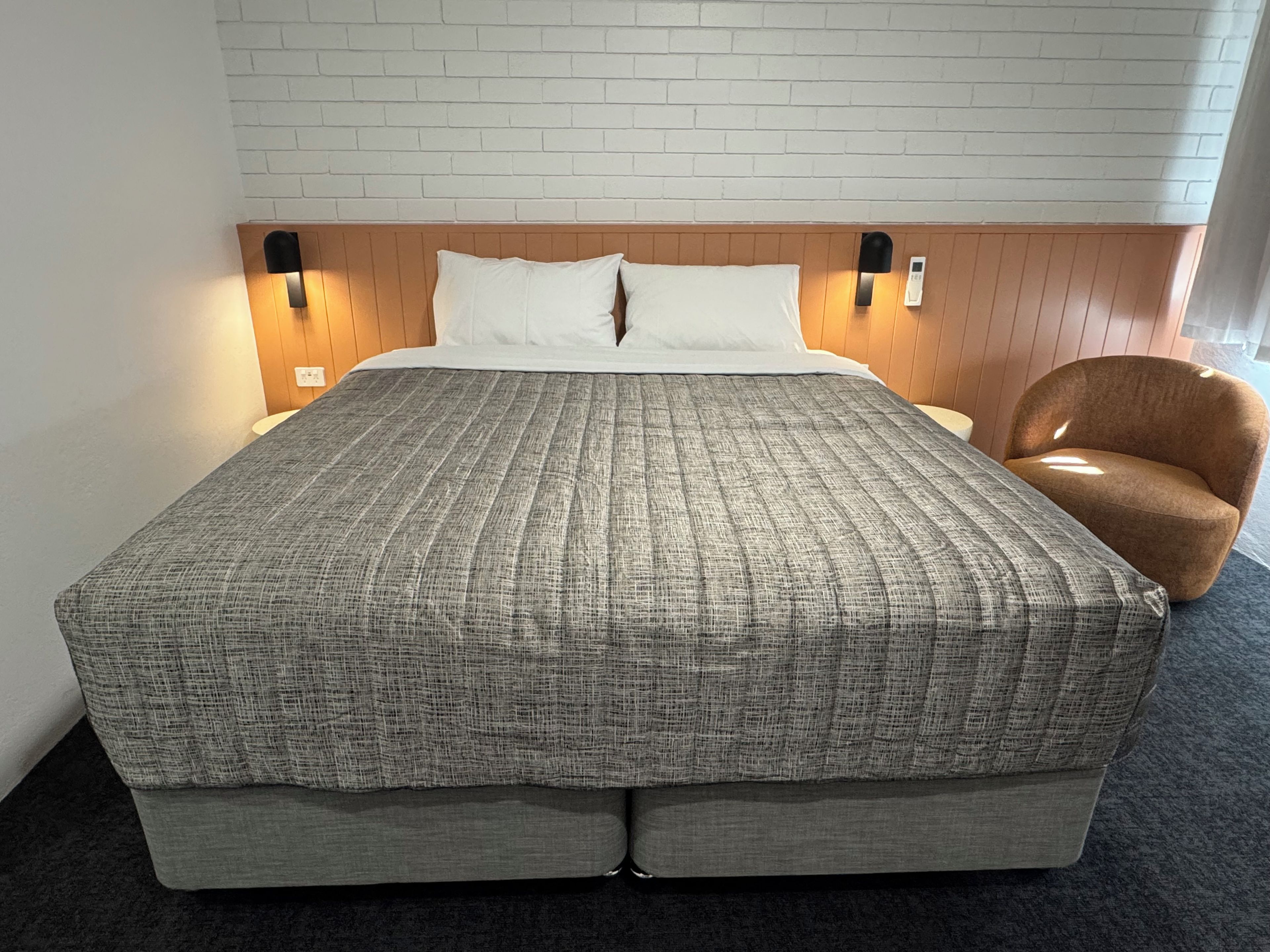 A hotel room with a large bed, a wooden headboard, two bedside lamps, and a brown armchair.