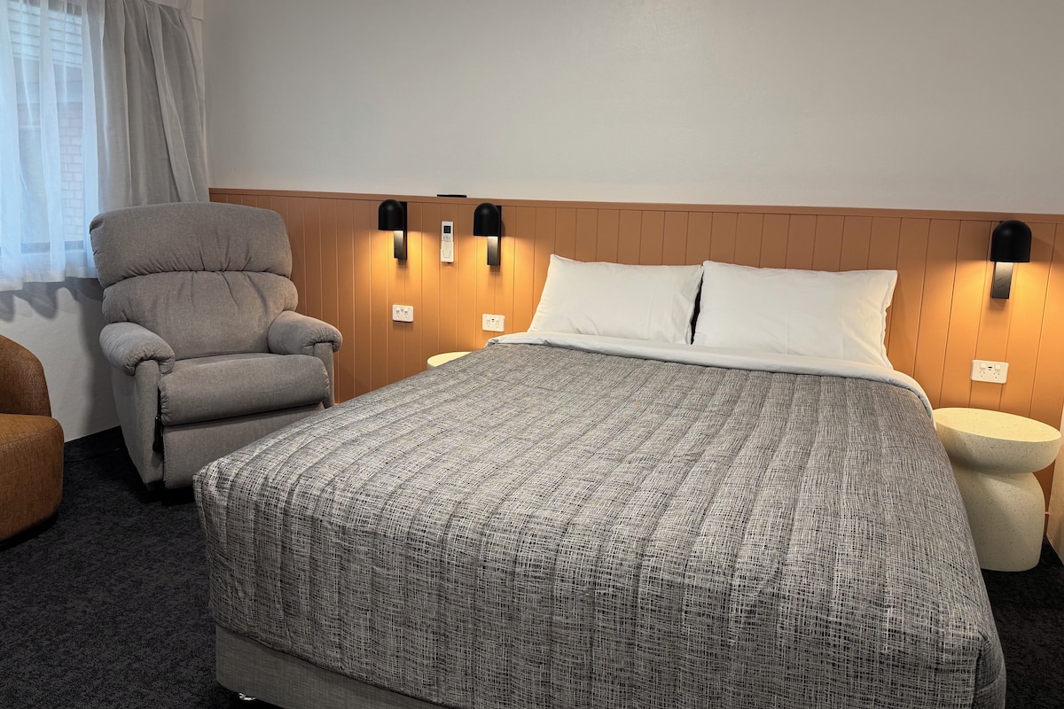 Habitación de hotel con una cama grande, un sillón gris, una mesita de noche redonda y una lámpara de pared.