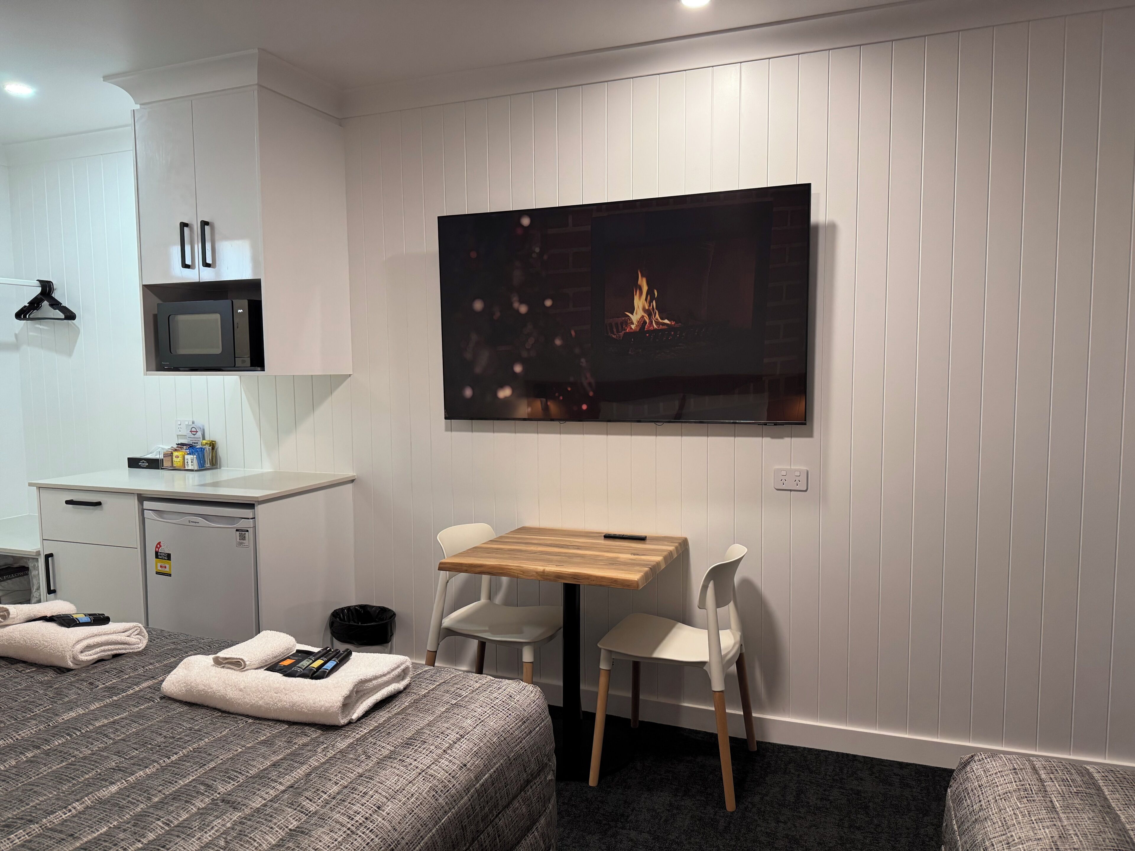 Habitación de hotel moderna con televisión de pantalla plana, una mesa de madera, dos sillas blancas, cama con ropa de cama gris, microondas y un mueble pequeño.