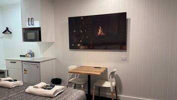 Habitación de hotel moderna con televisión de pantalla plana, una mesa de madera, dos sillas blancas, cama con ropa de cama gris, microondas y un mueble pequeño.